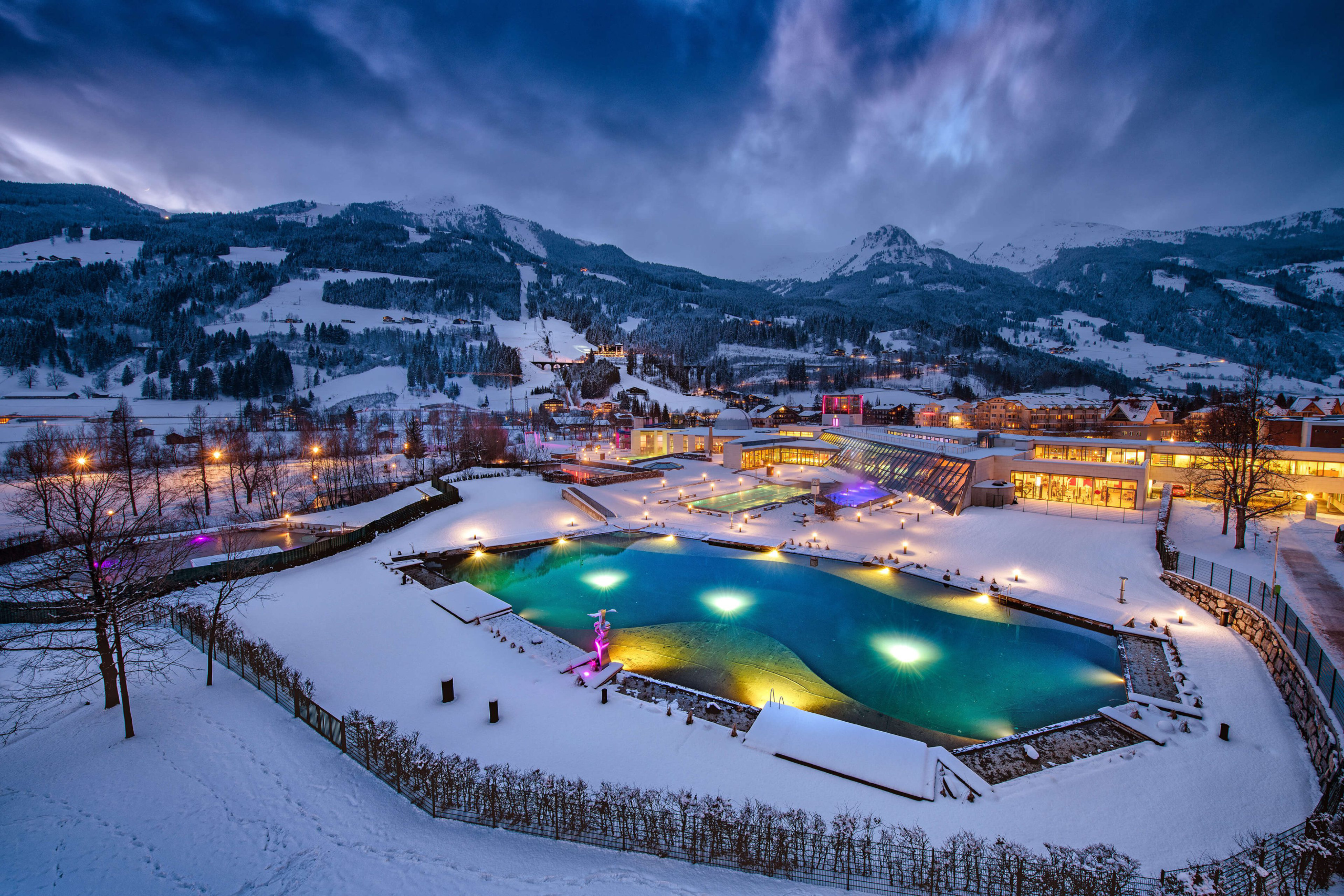 Winterliche Landschaft mit beleuchtetem Schwimmbecken und schneebedeckten Bergen.