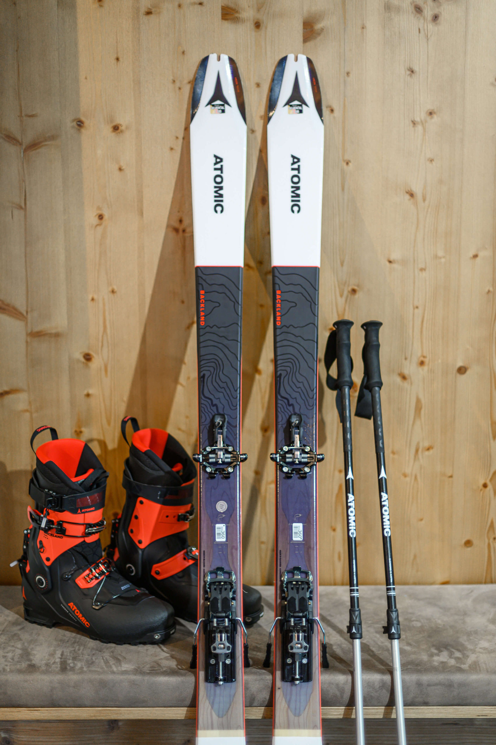 Skiausrüstung mit Atomic Skiern, Skischuhen und Stöcken vor rustikaler Holzwand.