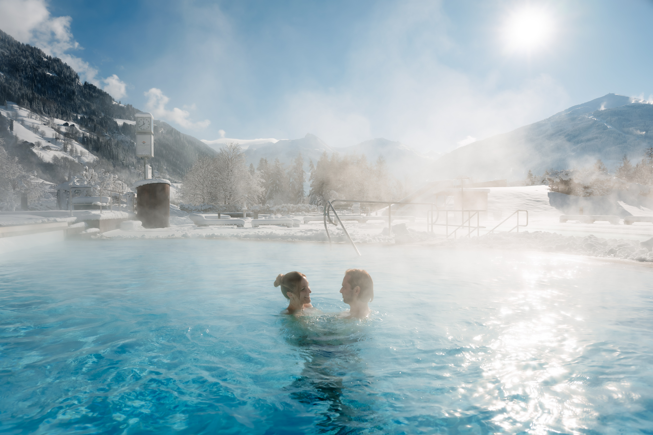 Zwei Personen im dampfenden Außenbecken der Alpentherme Gastein vor verschneiter Bergkulisse