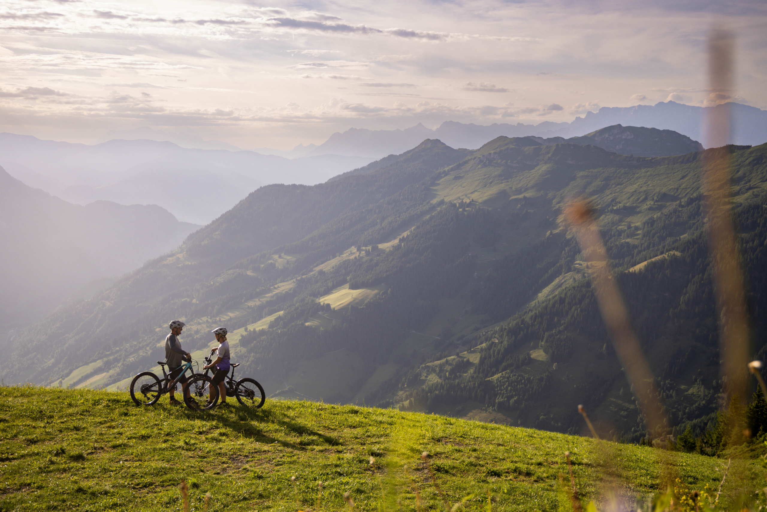 Zwei Mountainbiker stehen mit ihren Rädern auf einer grünen Bergwiese vor einem weiten Gebirgspanorama