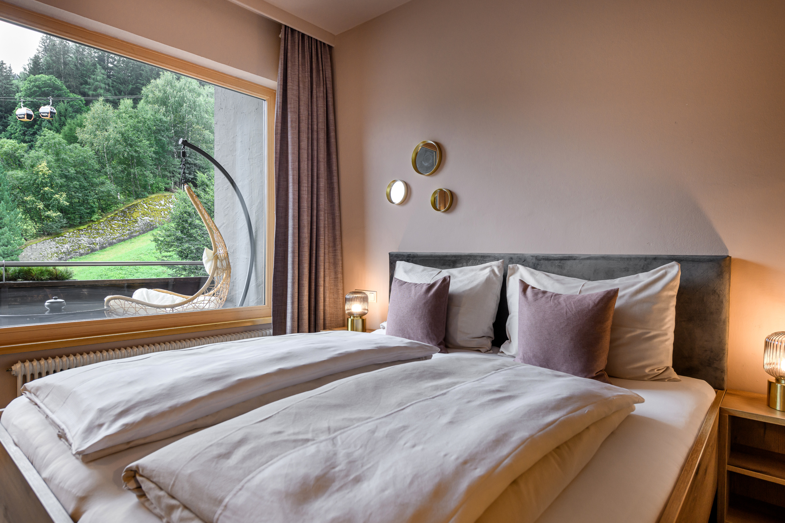 Doppelbett mit grauem Kopfteil, Nachttischlampen und Fensterblick auf bewaldeten Hang und Seilbahn