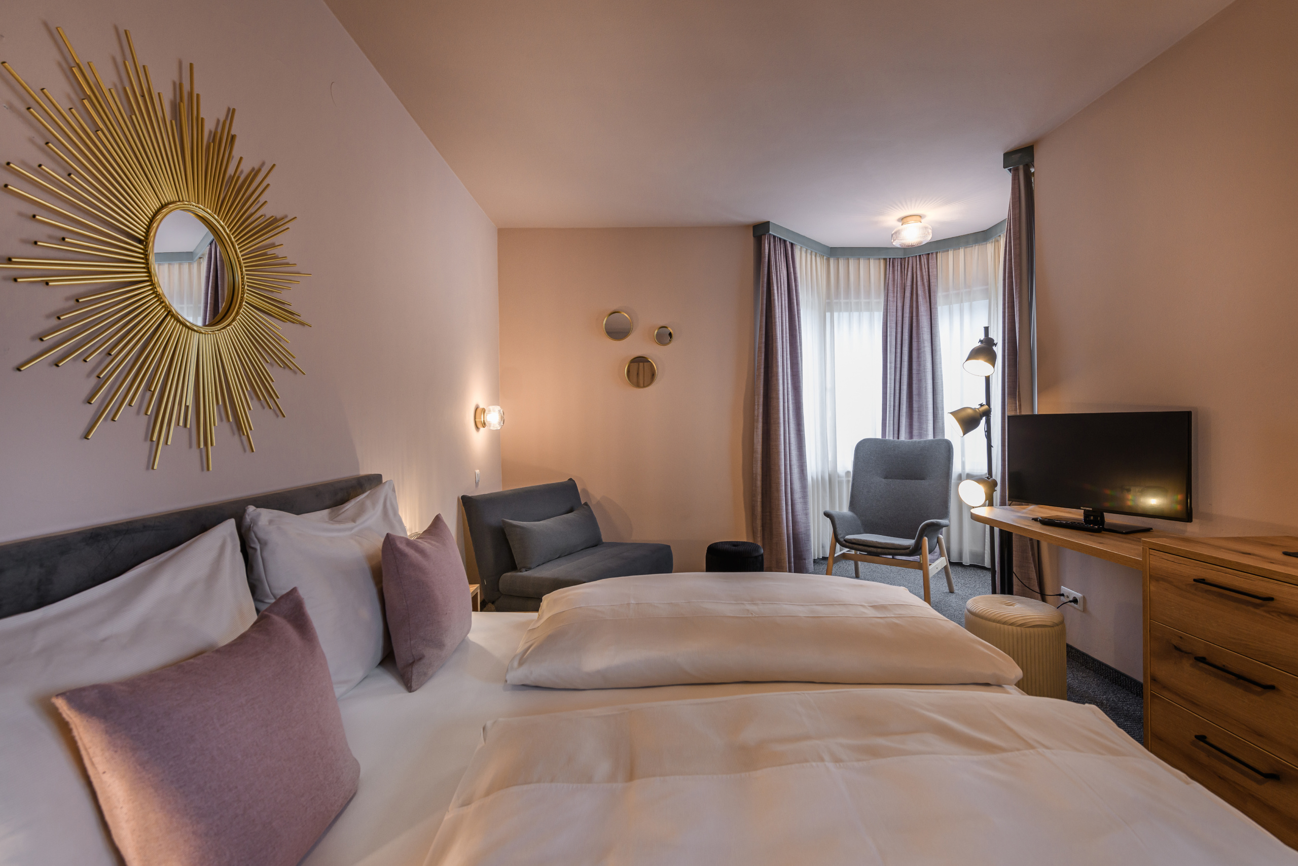 Doppelzimmer mit Doppelbett, Sesseln, Fernseher und großem goldfarbenem Sonnenspiegel an der Wand
