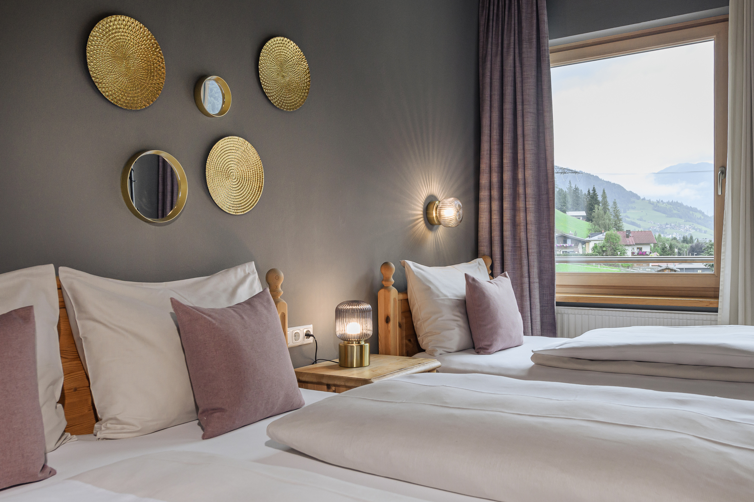 Dreibettzimmer mit zwei Holzbetten, grauer Akzentwand und Fenster mit Blick auf grüne Berge