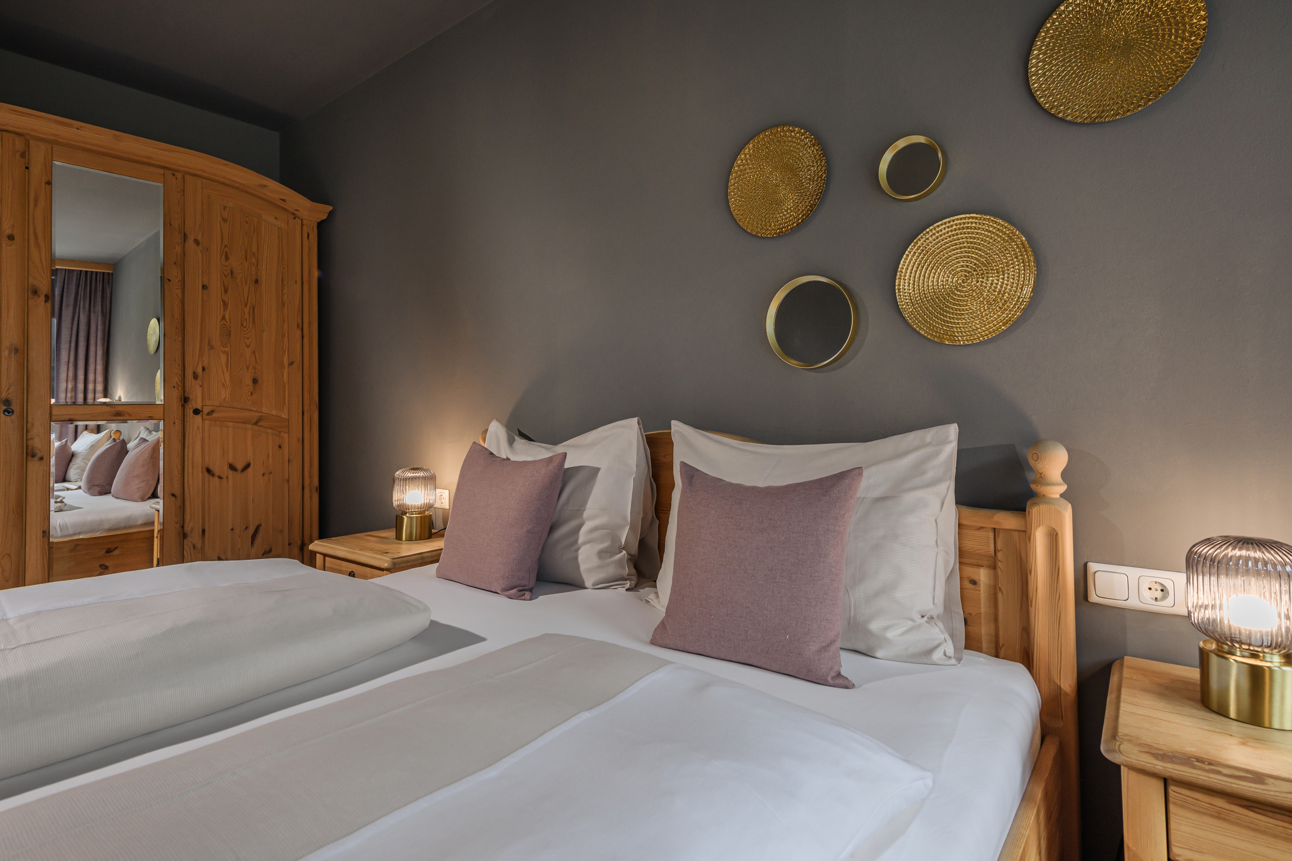 Doppelbett mit weißen Kissen und rosafarbenen Dekokissen in einem Zimmer mit Holzmöbeln und grauer Akzentwand