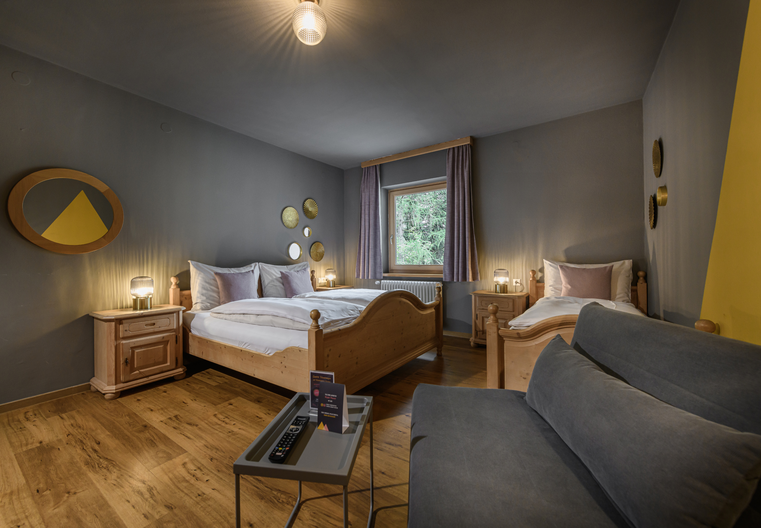 Vierbettzimmer mit zwei Holzbettgestellen, grauer Schlafcouch, Nachttischen und Fenster mit Vorhängen