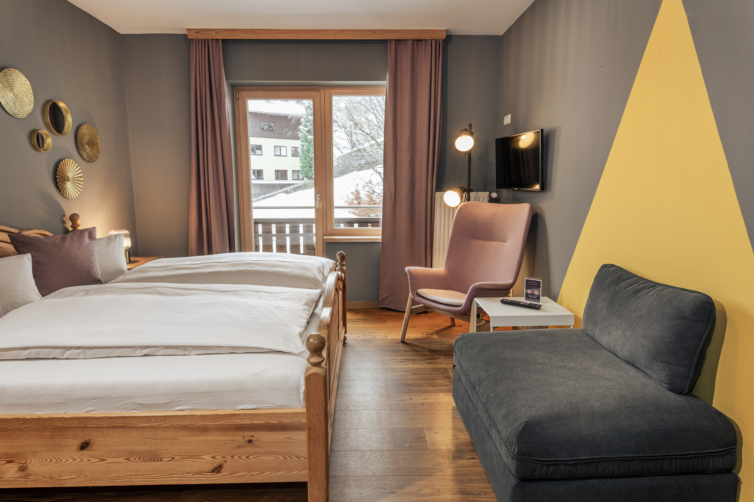 Doppelzimmer mit Holzbett, grauer Schlafcouch, Sessel und Balkon vor verschneiter Bergkulisse