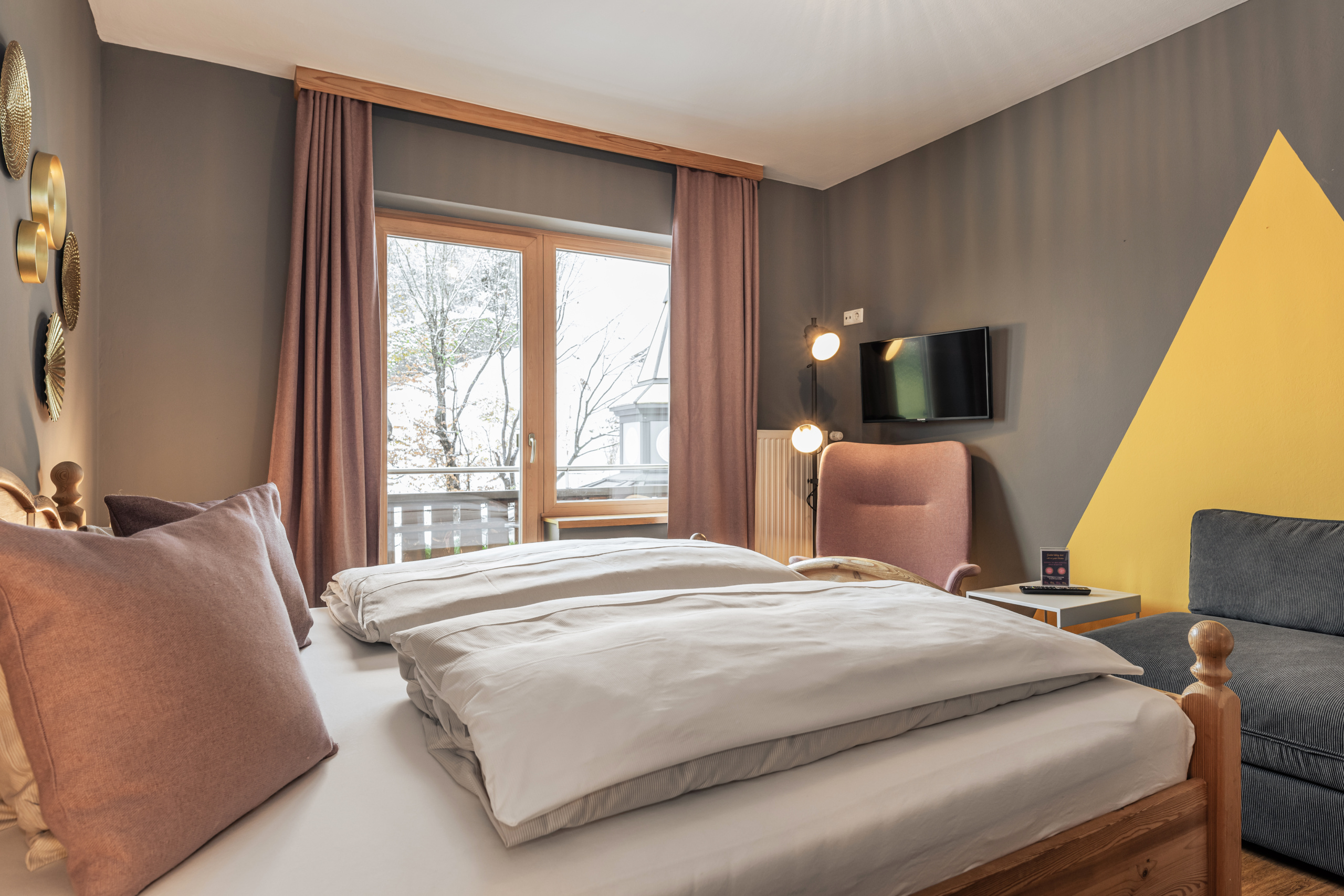 Doppelzimmer mit Holzbett, Schlafsofa, Sessel und Balkon vor winterlichen Bäumen