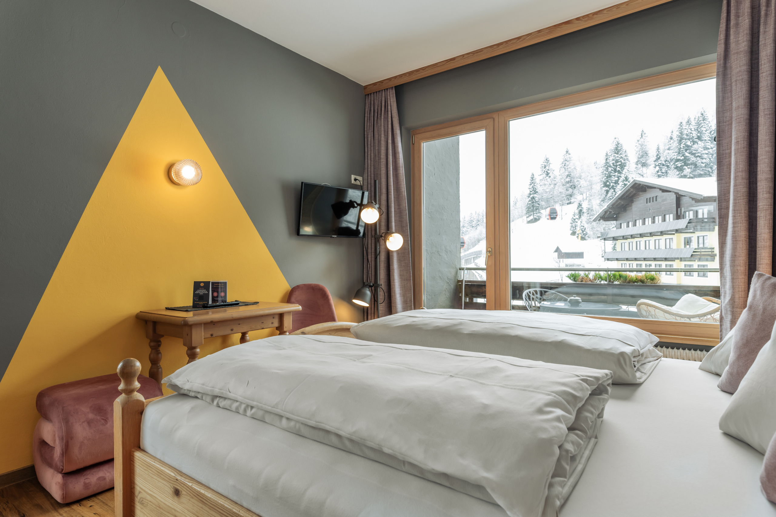 Doppelzimmer mit Holzbett, gelber Wandgestaltung und Balkonblick auf verschneite Berge