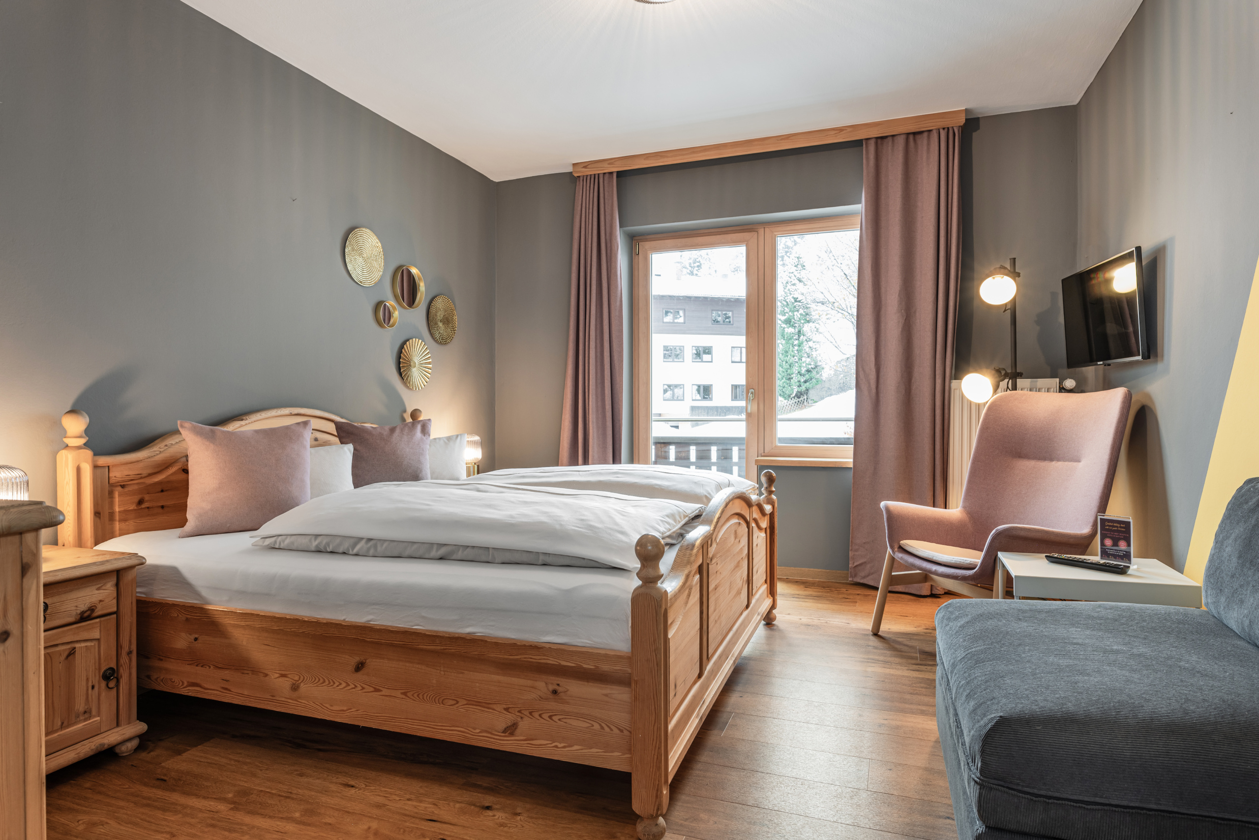 Doppelzimmer mit Holzbett, grauem Sofa, rosa Sessel und Fenster mit Blick auf Nachbarhäuser