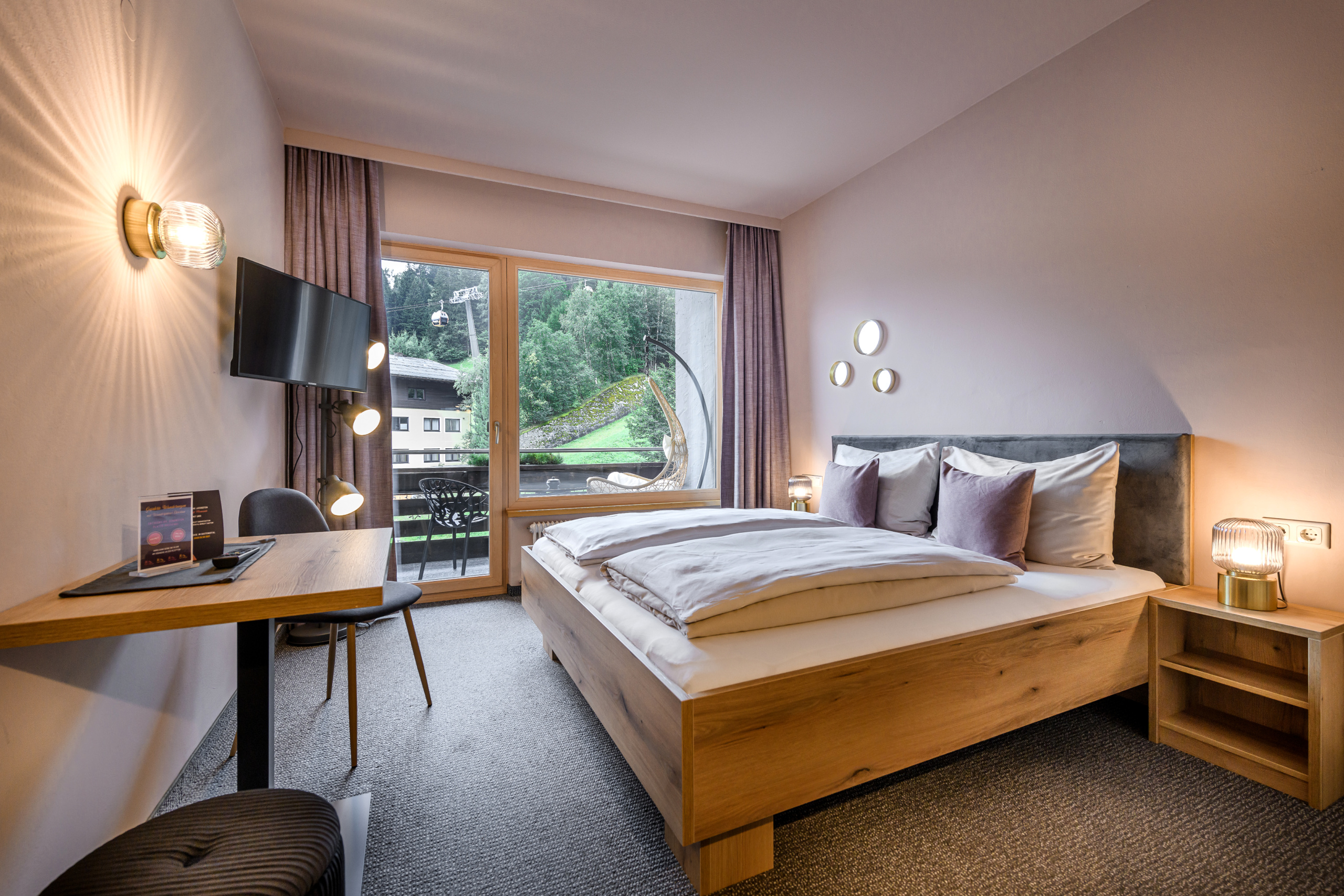 Helles Doppelzimmer mit Holzbett, Schreibtisch und Balkon mit Blick auf bewaldeten Hang