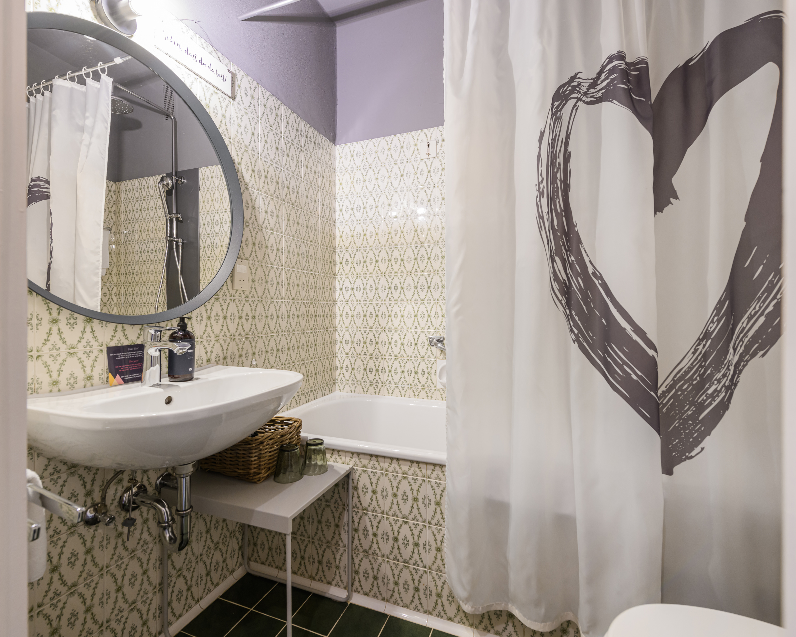 Badezimmer mit Waschbecken, runden Spiegel, Badewanne und weißem Duschvorhang mit Herzmotiv
