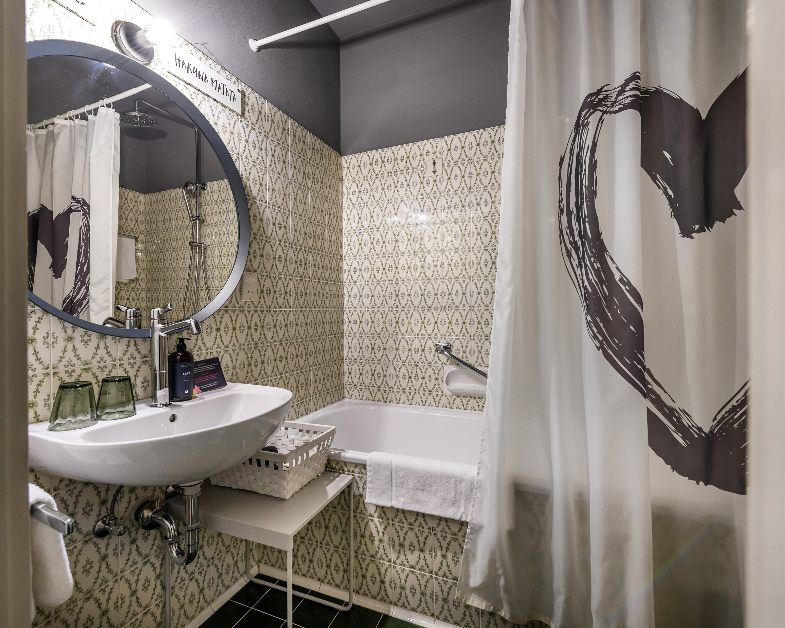 Badezimmer mit Waschbecken, Badewanne und Duschvorhang vor grün gemusterten Fliesen