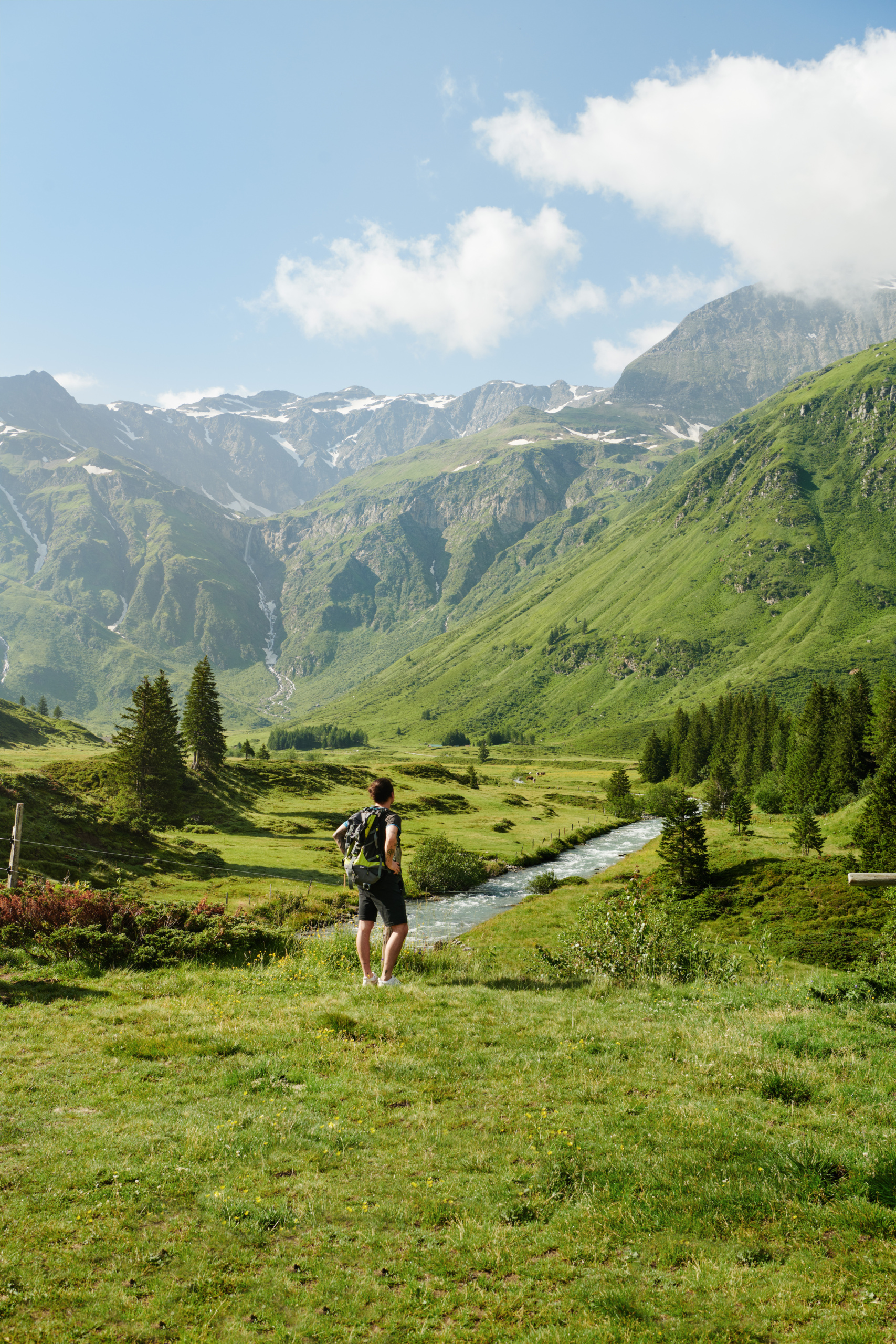 Wanderer mit Rucksack blickt über grünes Bergtal mit Bach und schneebedeckten Gipfeln