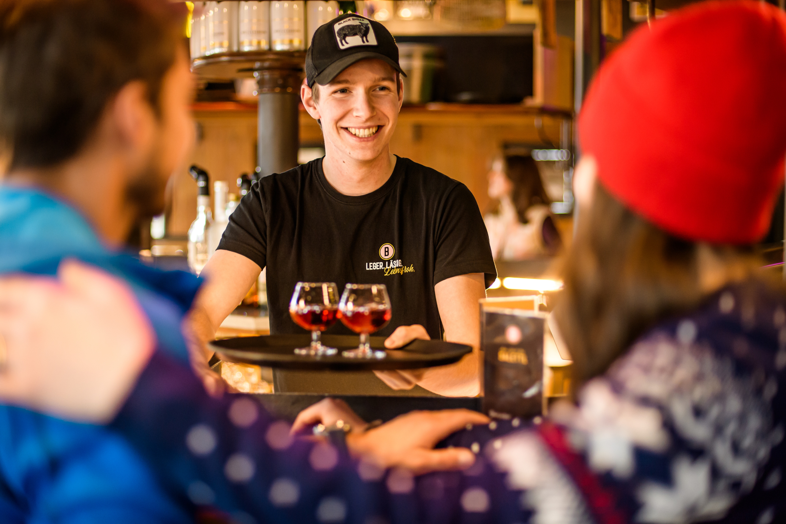 Lächelnder Kellner serviert zwei Gläser Rotwein an Gäste in einer gemütlichen Bar