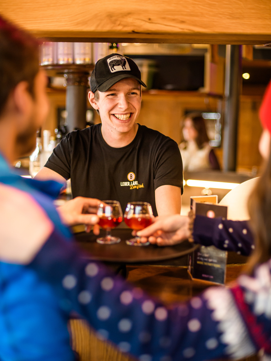 Lachender Barkeeper serviert zwei Gästen an der Holztheke je ein Glas Aperitif