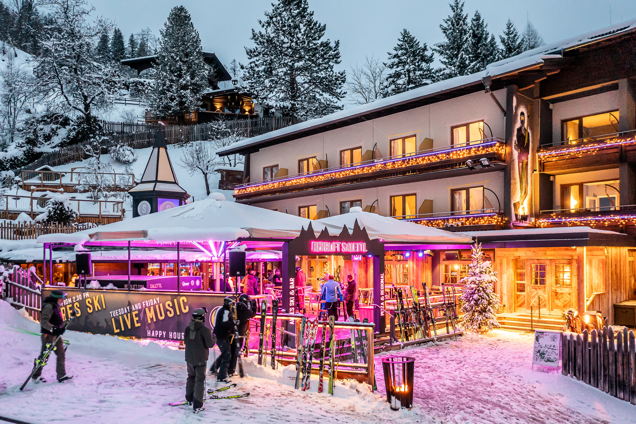 Beleuchtete Après-Ski-Bar mit Gästen, Skiern und verschneitem Hotelgebäude in der Dämmerung