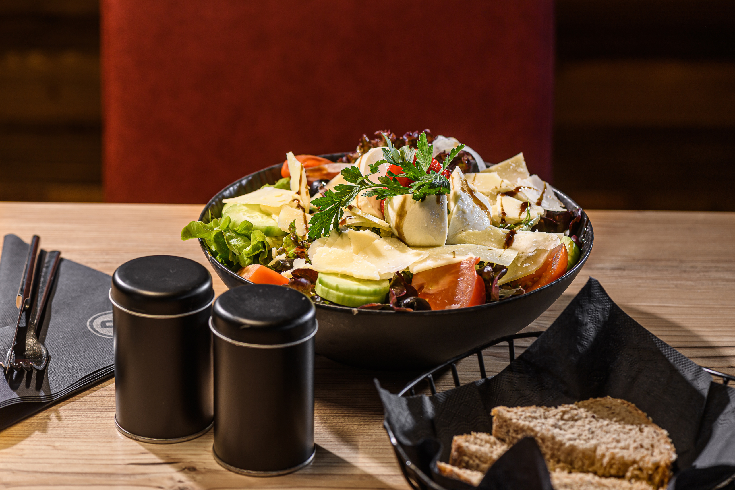 Großer gemischter Salat mit Käse, Gemüse und Kräutern auf einem Holztisch, dazu Brot und Gewürzdosen