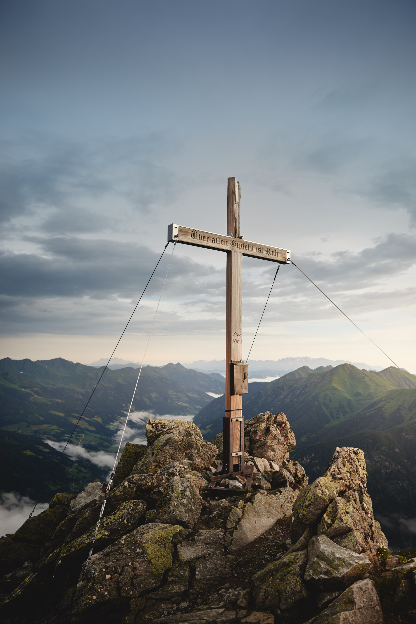 Hölzernes Gipfelkreuz auf felsigem Berggipfel mit Blick auf grüne Alpengipfel und wolkenverhangenes Tal