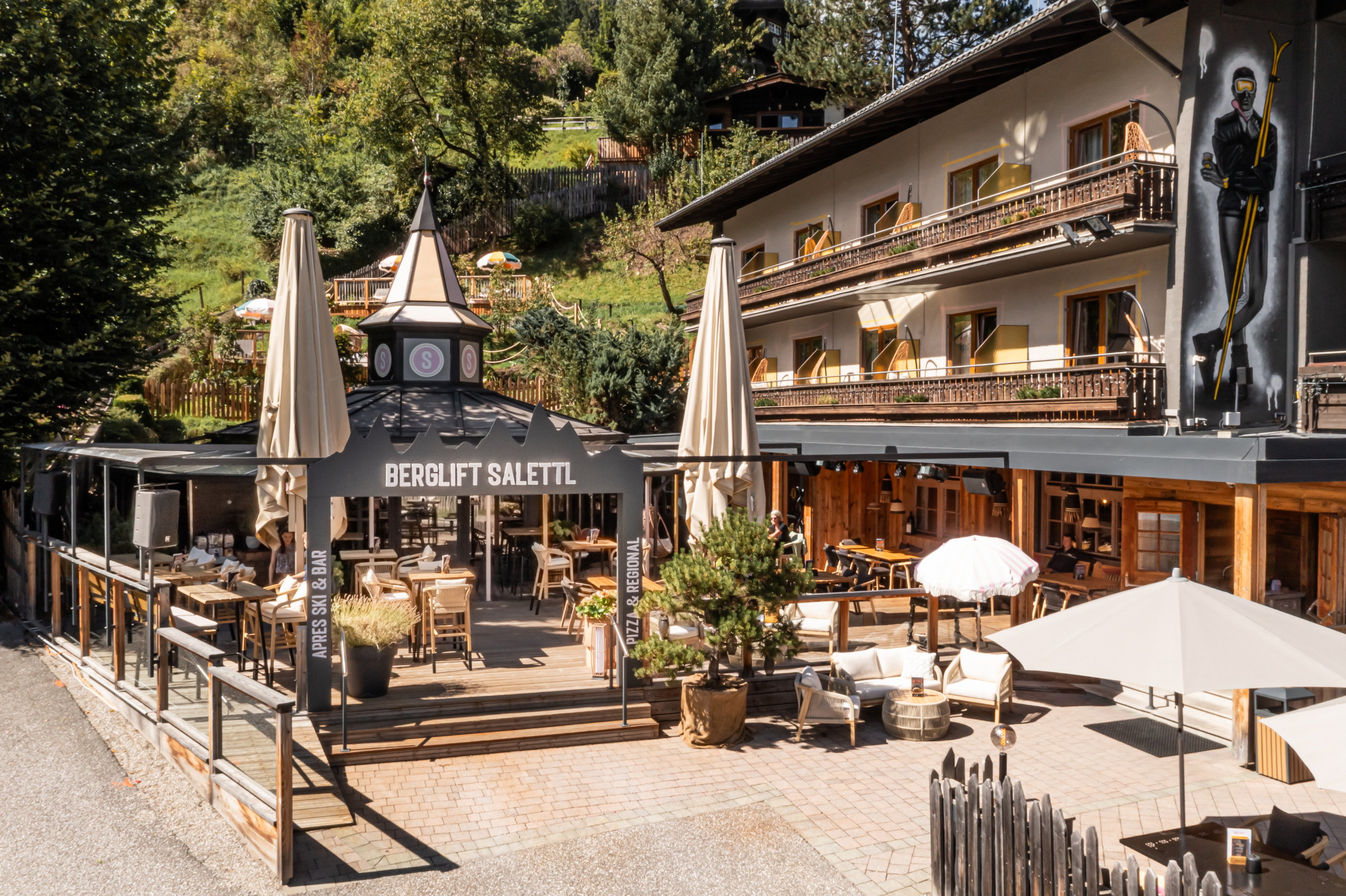 Sonnenterrasse des Berglift Salettl vor einem Hotel mit Balkonen, umgeben von Bäumen am Hang