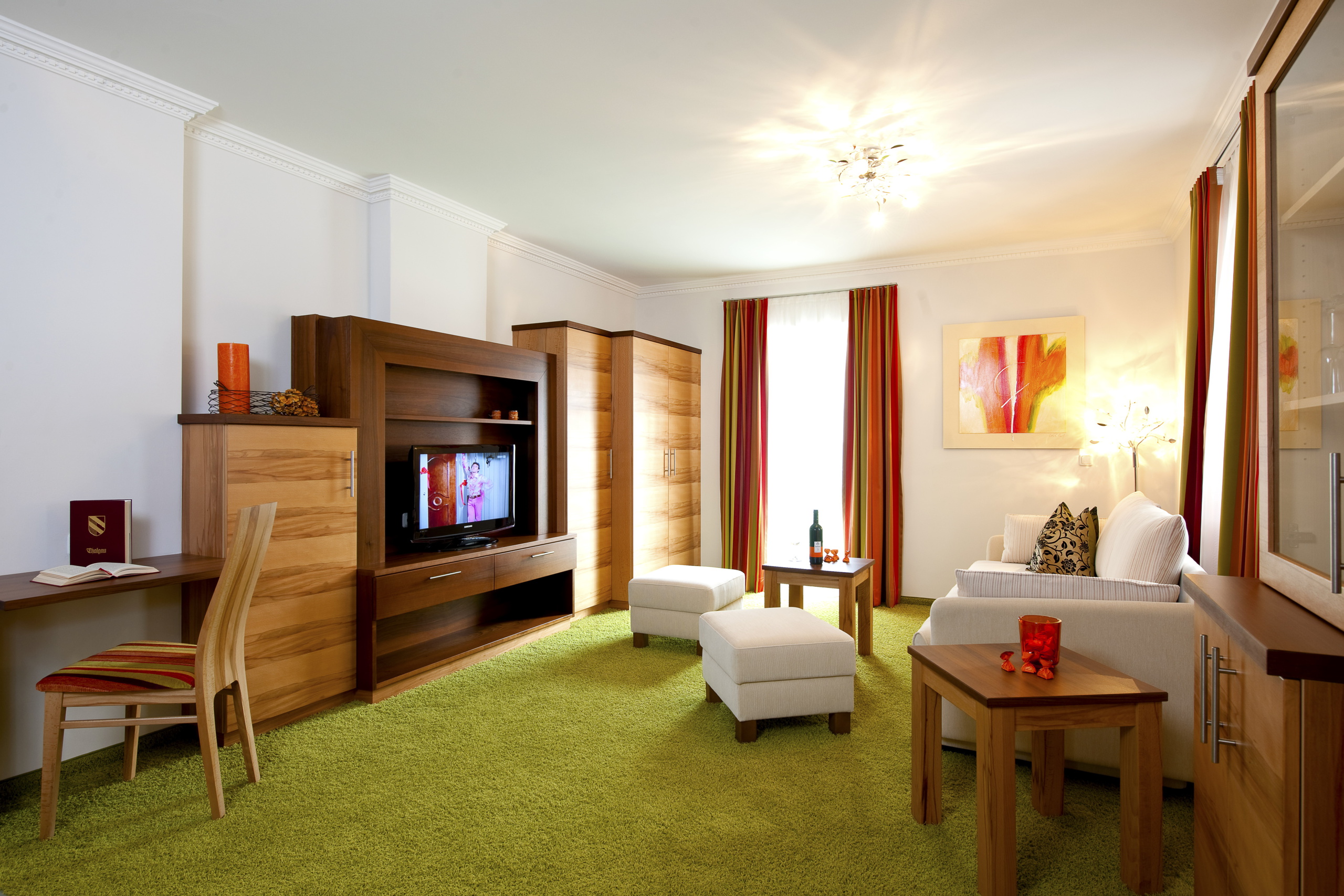 Heller Wohnbereich mit grüner Teppichfläche, Holzmöbeln, Sofa, Sesseln und gestreiften Vorhängen
