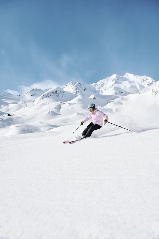 Skifahrerin in rosa Jacke fährt vor schneebedeckten Bergen durch tiefen Schnee